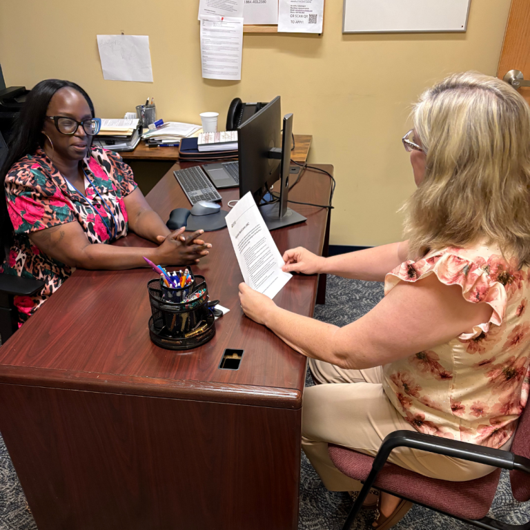 Two women are sitting across from each other at a desk, one is holding a paper while they have a conversation 