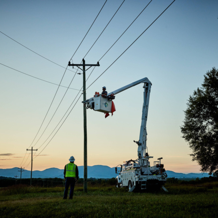 The image depicts an early morning where two men are working, both in reflective green vests, one standing on the grass while the other in a lift going up to fix lines