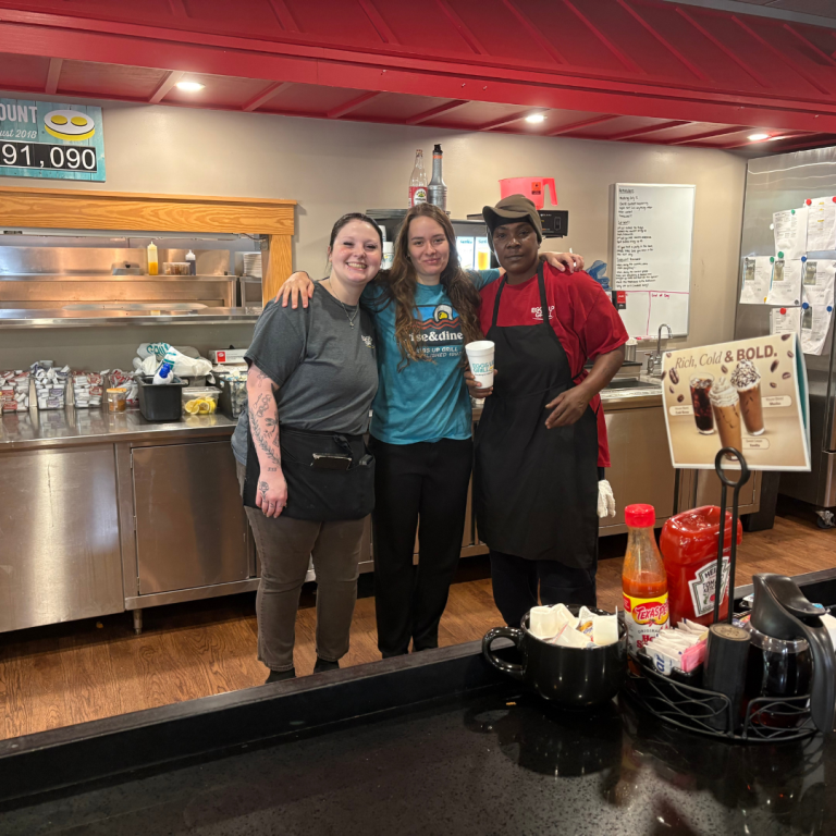 Three workers at Eggs Up Grill are are standing together behind the counter, smiling and posing for the picture 