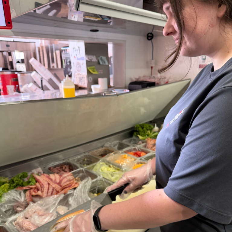 A woman is standing in a sub shop, behind the counter, in front of all the toppings and sub add-ons, making a sandwich