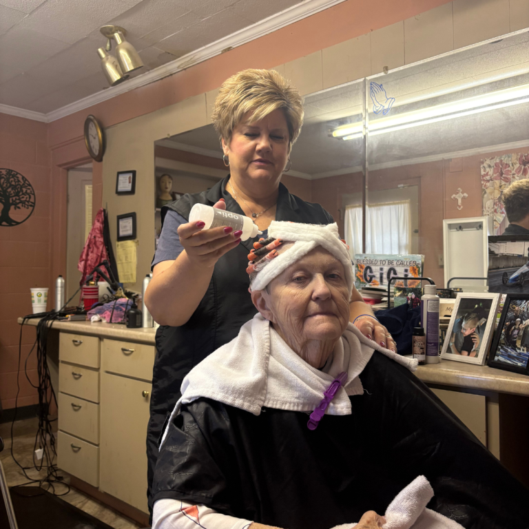 Two women are in a hair salon. One lady is behind the other, putting material in the women's hair. The older lady, is sitting in the chair, with a towel around her neck, and rollers in her hair