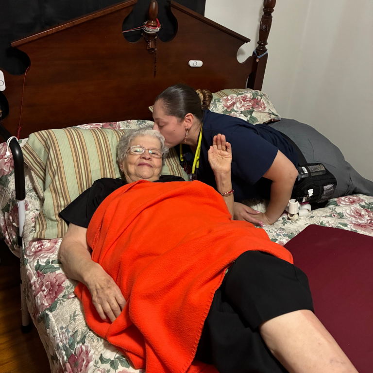 Two ladies appear in the shot. One lady, on the edge of the bed is leaned over, kissing the other lady on the side of the head, an elderly lady laying on the bed. The older lady is waving at the camera with a red blanket over her