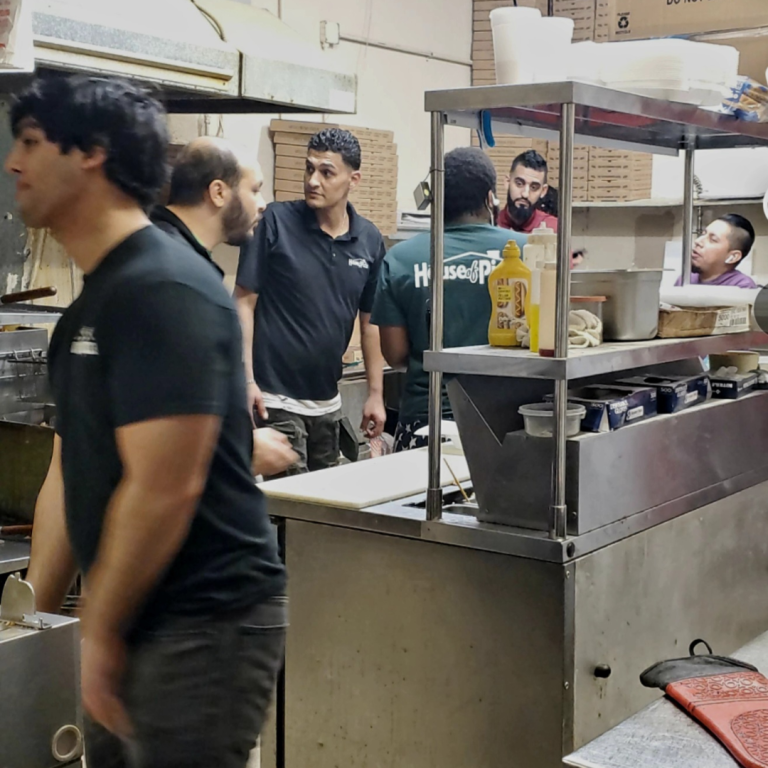 Several men gather around a kitchen working and making pizza 