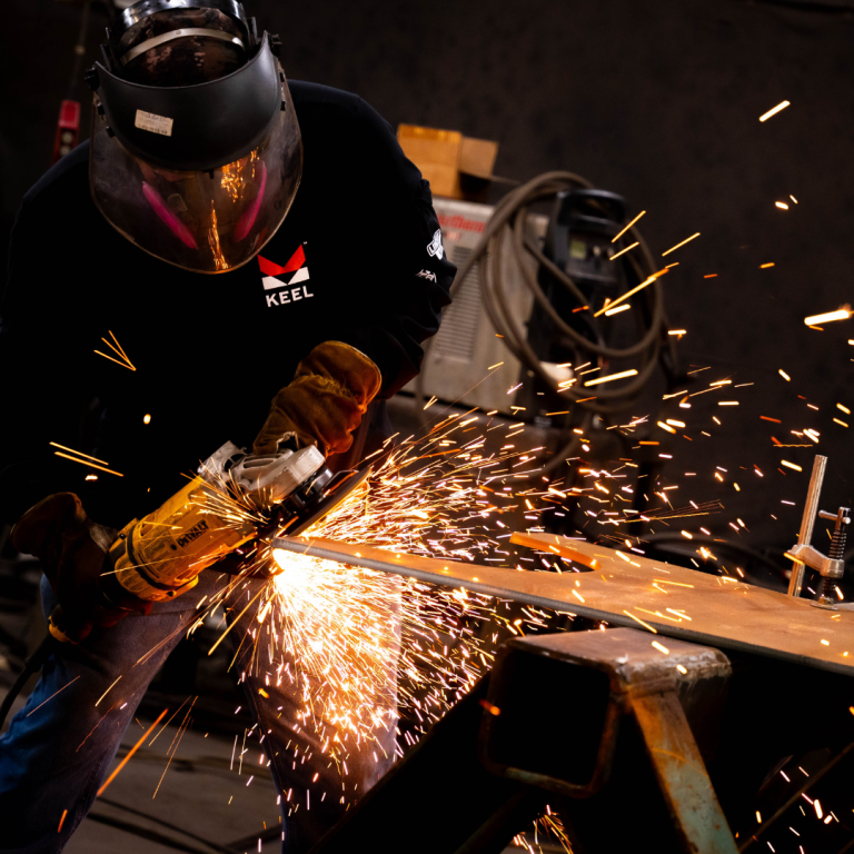 A man stands in full protective clothes with the Keel insignia, sawing with an electric saw at a piece of metal, sparks flying 