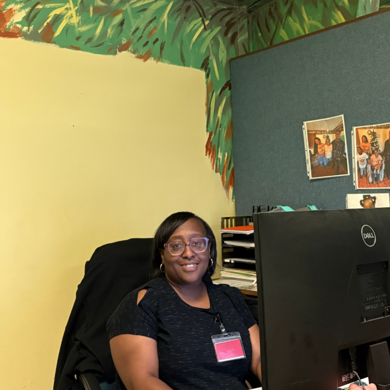 A woman sits at her desk in her office in front of a computer, doing work. She looks and smiles at the camera