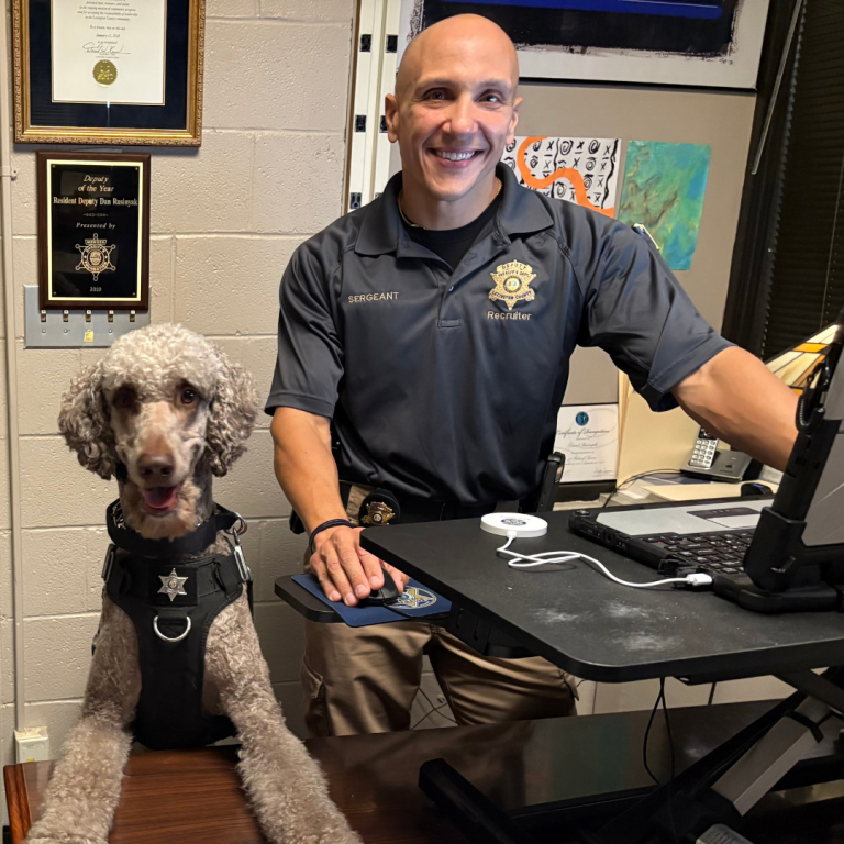 A man stands behind his desk, working on his computer. A police dog stands on his hind legs, the dogs feet on the desk. Both the dog, and the police officer are smiling at the camera 