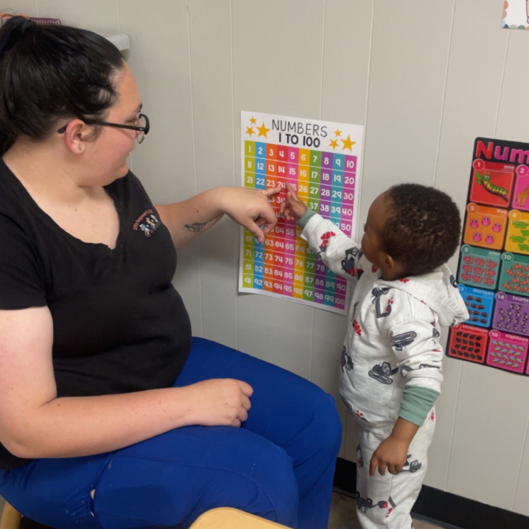 A woman is sitting on a chair next to a little boy, they are both pointing at and counting a poster on the wall with numbers 1-100