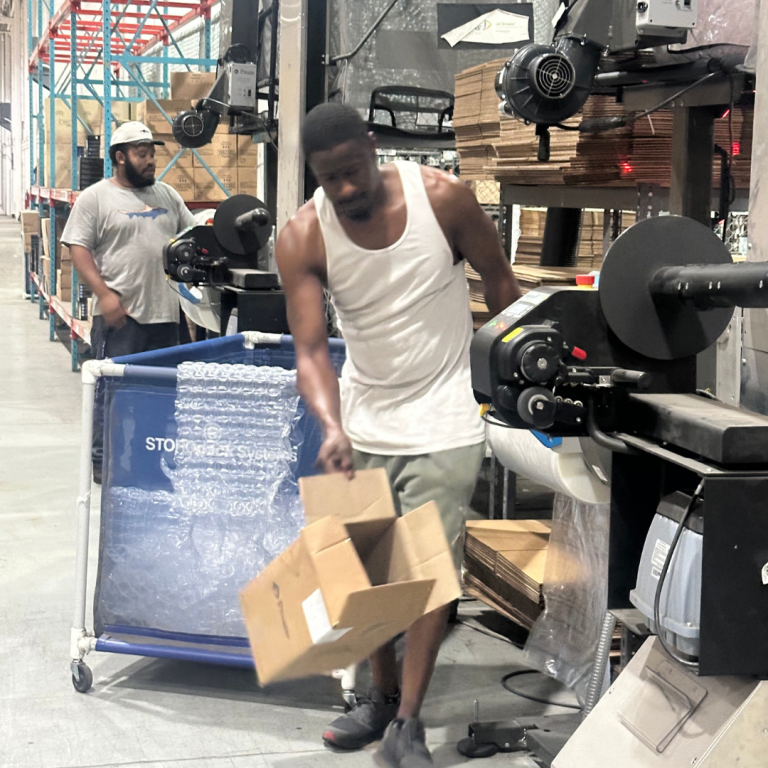 A man in a warehouse, working, carries two carboard boxes
