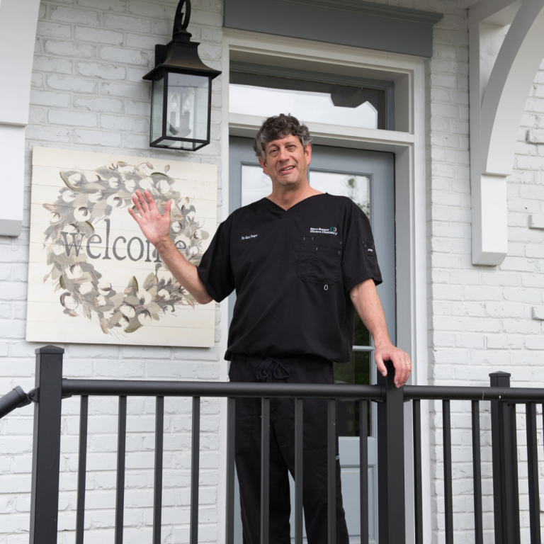 A man is standing outside of a building, next to a sign that says 'welcome' he is smiling and waving at the camera