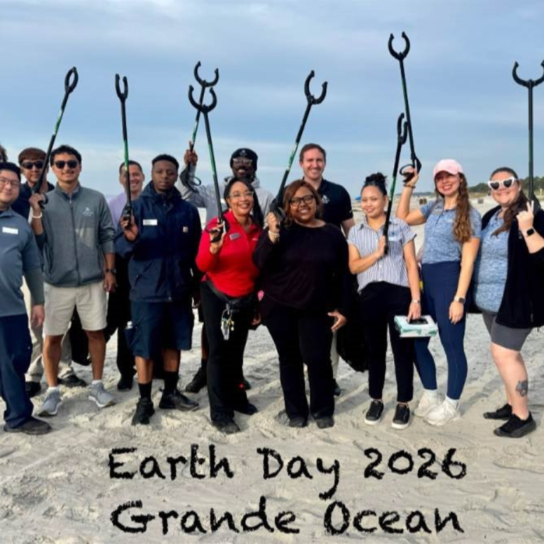 A group of people stand on the sand looking at the camera and smiling. They are all holding trash grabbers