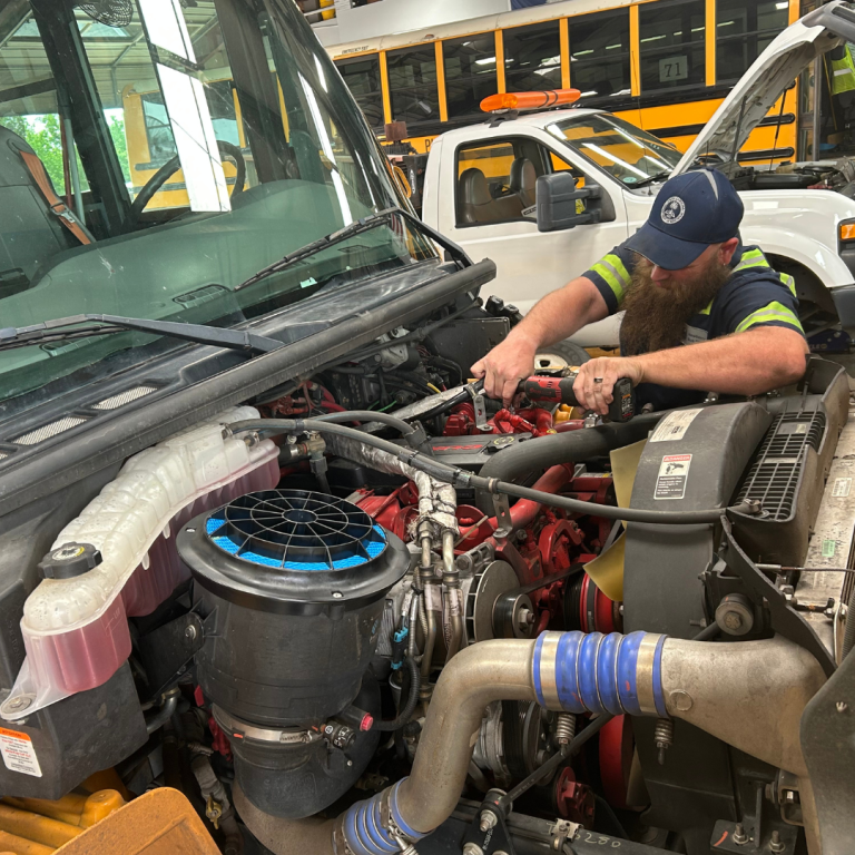 A man works on the front of a bus, with the hood off, he uses a tool to work on the bus