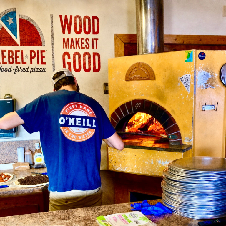 A man facing away, wearing a Rebel Pie shirt, puts a pizza into a pizza oven 