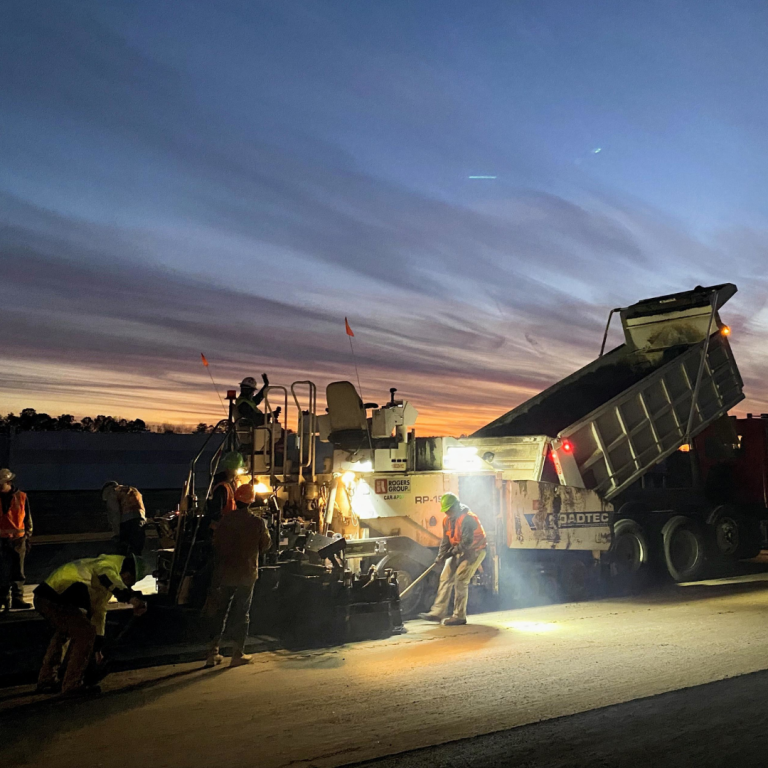 Several construction workers work around the side of a road with a large dump truck 